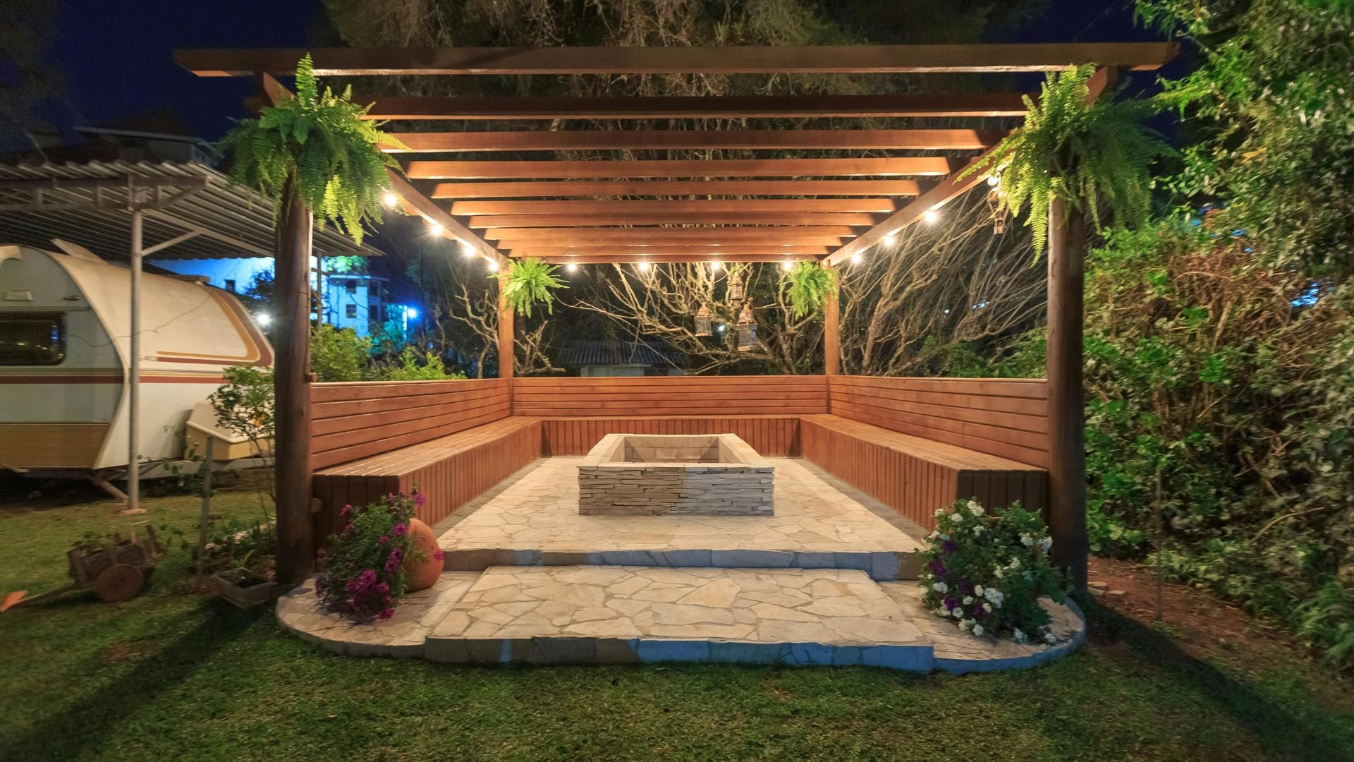 Wooden pergola with stone firepit, string lights, and hanging ferns at night