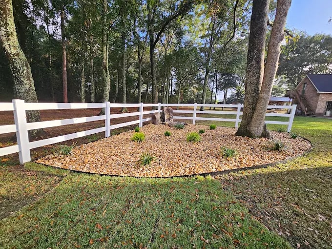 Circular mulch bed with small plants, white fence, and trees in background