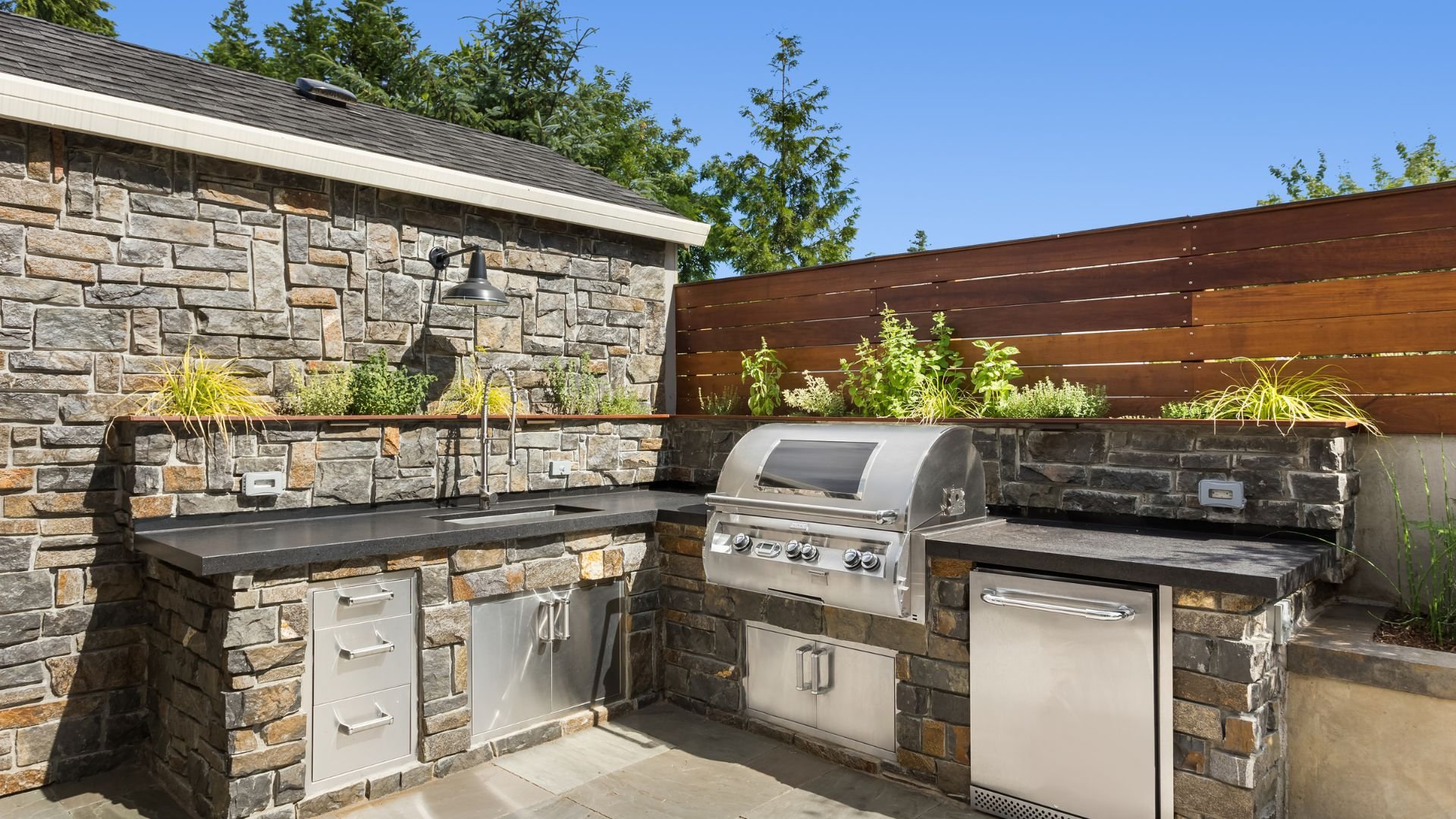 Outdoor kitchen with stone walls, grill, and wooden fence surrounded by plants